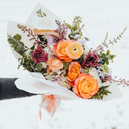 Hand-tied bouquet of orange and yellow blooms with burgundy accents wrapped in white paper.