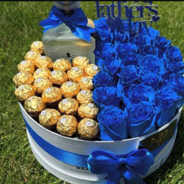Blue roses and gold-wrapped chocolates arranged in a round gift box with a ribbon.