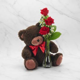 Teddy bear beside a small vase of red roses and white daisies