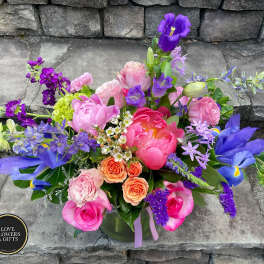 Vibrant mixed arrangement of pink peonies, roses, and purple irises in a clear glass vase