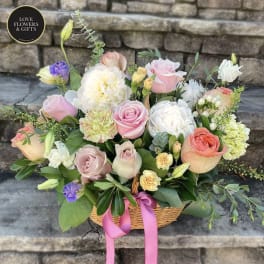 Basket arrangement of pink roses, white peonies, and pastel blooms with a pink ribbon