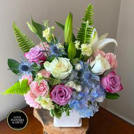 Pastel roses, lilies, and blue hydrangea in a white square vase with ferns.
