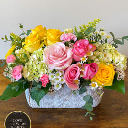 Low arrangement of pink and yellow roses with hydrangeas and daisies in a rectangular white container