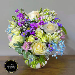 Round glass vase of cream roses, purple blooms, and blue accents in a mixed floral arrangement