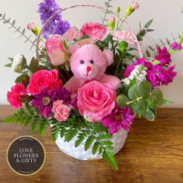 White basket of pink and purple flowers with a pink teddy bear in the center
