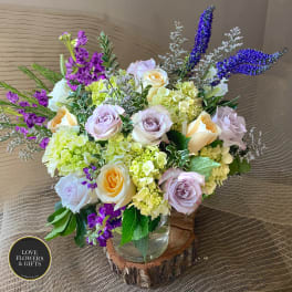 Arrangement of cream and lavender roses with green hydrangeas and purple flowers in a clear vase on a wood slice.