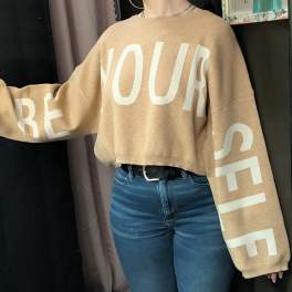 Person wearing a tan sweater with large white lettering and blue jeans