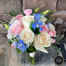 Hand-tied bouquet of ivory and pink roses with blue delphinium, wrapped in white ribbon against a stone background.