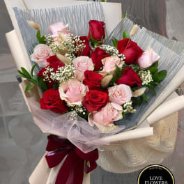 Hand-tied bouquet of red and pink roses with white filler flowers wrapped in cream and gray paper.