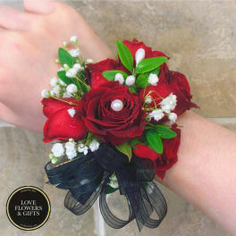 Red rose wrist corsage with white filler flowers, pearls, and a black ribbon bow on a wrist