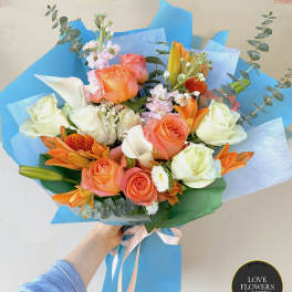 Hand-tied bouquet of peach and white roses with orange lilies and white blooms in blue wrapping.