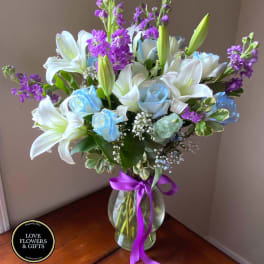Tall vase arrangement with white lilies, blue roses, and purple flowers tied with a purple ribbon