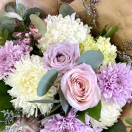 Hand-tied bouquet of pastel pink and lavender roses with white and lavender chrysanthemums in kraft paper.