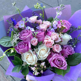 Hand-tied bouquet of lavender, pink, and white roses with accent flowers wrapped in purple paper.