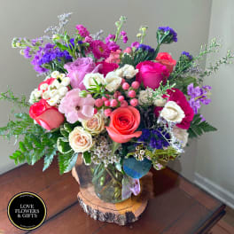 Colorful mixed bouquet of roses and assorted blooms in a clear glass vase on a wood slice.