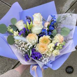 Hand-tied bouquet of cream roses, blue delphinium, and mixed pastel flowers wrapped in lavender paper.