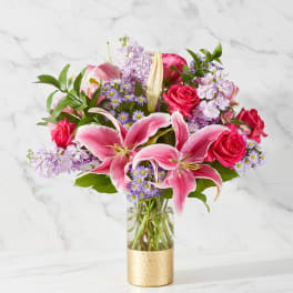 Pink lilies and roses arranged in a glass vase with purple filler flowers
