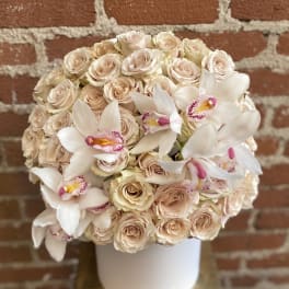 Round hatbox arrangement of blush roses with white orchids against a brick wall.
