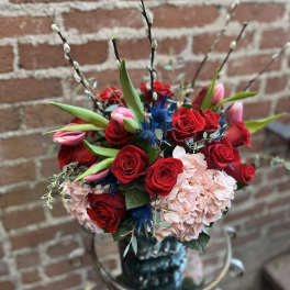 Tall arrangement of red roses, pink tulips, and pink hydrangea with blue accent flowers in a dark vase.