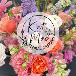 Mixed bouquet of pink, peach, and purple flowers with roses and delphinium, with a circular florist logo overlay.