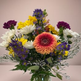 Mixed bouquet in a glass vase with purple, yellow, white, and peach flowers.