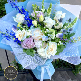 Hand-tied bouquet of white roses, blue delphinium, and pastel blooms wrapped in blue paper.