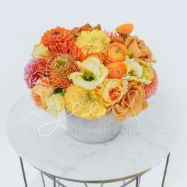 Low arrangement of orange, yellow, and peach flowers in a white pot on a round marble table.