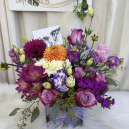 Mixed bouquet of purple, pink, orange, and white flowers in a square vase with ribbon.