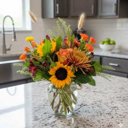 Mixed bouquet of sunflowers, roses, and orange blooms in a glass vase