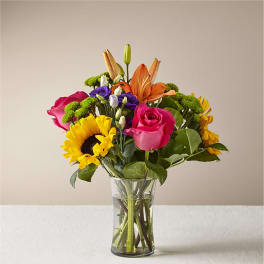Mixed bouquet of roses, lilies, sunflowers, and purple blooms in a glass vase
