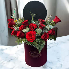 Red roses arranged in a burgundy hatbox with greenery