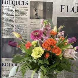 Mixed bouquet of orange, pink, purple, and yellow flowers in a clear glass vase