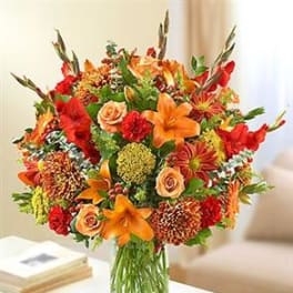 Tall mixed bouquet of orange lilies, peach roses, and red flowers in a clear glass vase