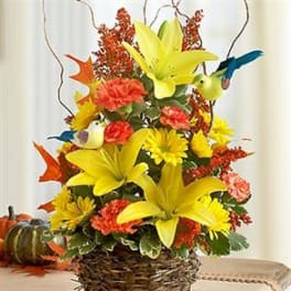Basket arrangement of yellow lilies, yellow daisies, orange carnations, and small decorative birds in autumn colors.