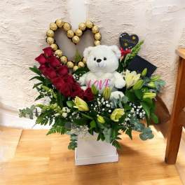 Red roses and white lilies with a teddy bear in a heart-shaped gift arrangement