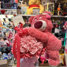 Pink teddy bear hugging a bouquet of pink roses in a glass vase