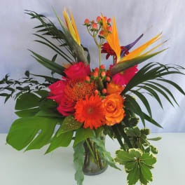 Tall tropical arrangement with orange gerberas, roses, and birds of paradise in a clear glass vase.