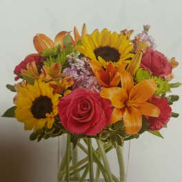 Mixed bouquet of sunflowers, roses, lilies, and other blooms in a clear glass vase