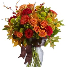 Autumn arrangement of orange roses, red carnations, green mums and fall leaves in a clear glass vase