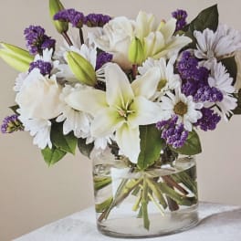 White lilies, daisies, and purple filler flowers in a clear glass vase