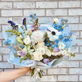 Handheld bouquet of white, blue, and lavender flowers wrapped in pale blue paper
