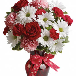 Red roses, white daisies, and pink carnations in a red vase with a ribbon