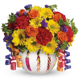 Bright mixed bouquet of yellow daisies, orange roses and red carnations in a white candle-print bowl with curling ribbons.