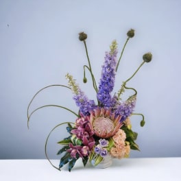 Tall modern arrangement with purple delphinium, a large protea, orchids, and peach blooms in a white vase