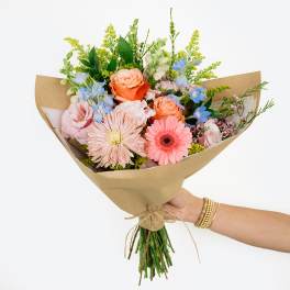 Hand-tied bouquet of pink, peach, and blue flowers wrapped in kraft paper