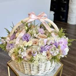 Pastel roses and orchids arranged in a white wicker basket with a pink ribbon