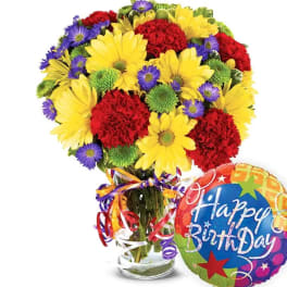 Bouquet of yellow daisies, red carnations, and purple asters in a glass vase with a birthday balloon