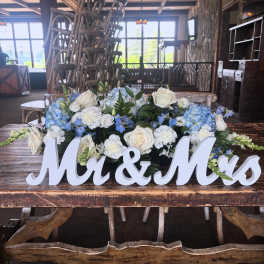 Low wedding centerpiece of white and blue flowers with a white Mr & Mrs sign on a rustic wooden table