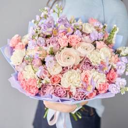 Large pastel bouquet of pink roses, white blooms, and lavender flowers wrapped in lilac paper, held in both hands
