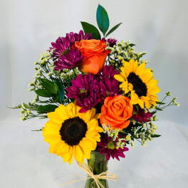 Bouquet of sunflowers, orange roses, and purple daisies in a glass vase